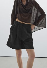 Personne portant un short ample noir plissé et un haut asymétrique à manches longues en tulle marron foncé, debout avec les mains dans les poches.