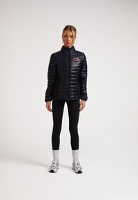 Marineblauer gesteppter Pufferjacke mit hohem Kragen und schwarzen Seitenpaneelen, mit einem Logo auf der Brust. Getragen mit schwarzen Leggings und Schuhen.