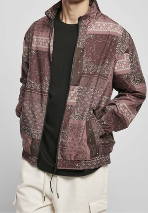 Personne portant une veste zippée à motifs bordeaux et marron sur une chemise noire et un pantalon blanc, les mains dans les poches de la veste.