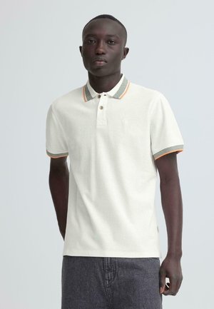 Jeune homme portant un polo à manches courtes blanc avec des bordures grises et orange, debout devant un fond gris clair uni.