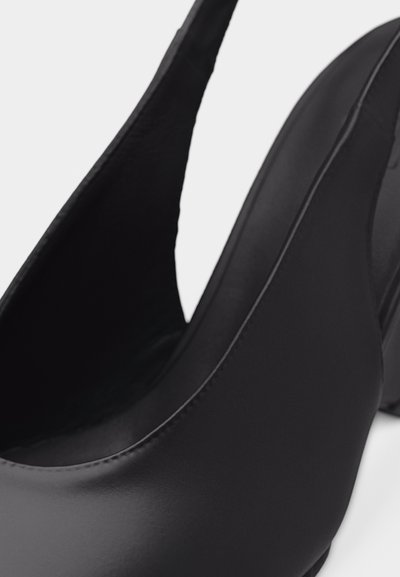 Μαύρο δερμάτινο παπούτσι με ψηλό τακούνι, μυτερή μύτη και ιμάντα τύπου slingback, εμφανίζεται σε κοντινό πλάνο πάνω σε μονόχρωμο φόντο.