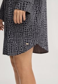 Dunkelgraues Kleid mit einem strukturierten, gitterartigen Muster in Schwarz. Verfügt über einen seitlichen Schlitz und ein kleines metallisches Logo-Detail nahe dem Saum.