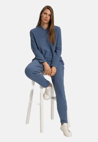 Felpa blu e pantaloni coordinati realizzati in morbido materiale, caratterizzati da una vestibilità comoda. Modello seduto su uno sgabello bianco, indossando sneaker bianche.