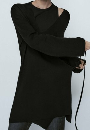 Person iført en løs sort asymmetrisk trøje med den ene skulder eksponeret, der holder en lang sort snor, mod en ensfarvet baggrund.