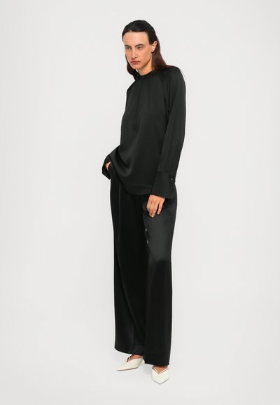 Woman wearing loose black satin blouse and wide pants with white pointed heels, standing against plain white background.