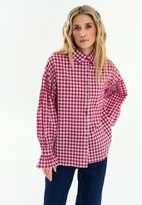 Žena s dlouhými vlnitými blond vlasy, oblečená v červenobílé kostkované blůze s volánkovými manžetami a tmavě modrých kalhotách, stojící na bílém pozadí.