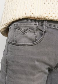 Jeans de denim gris con un bolsillo trasero de diseño distintivo, textura sutil y un corte ajustado. La tela parece suave y envejecida.