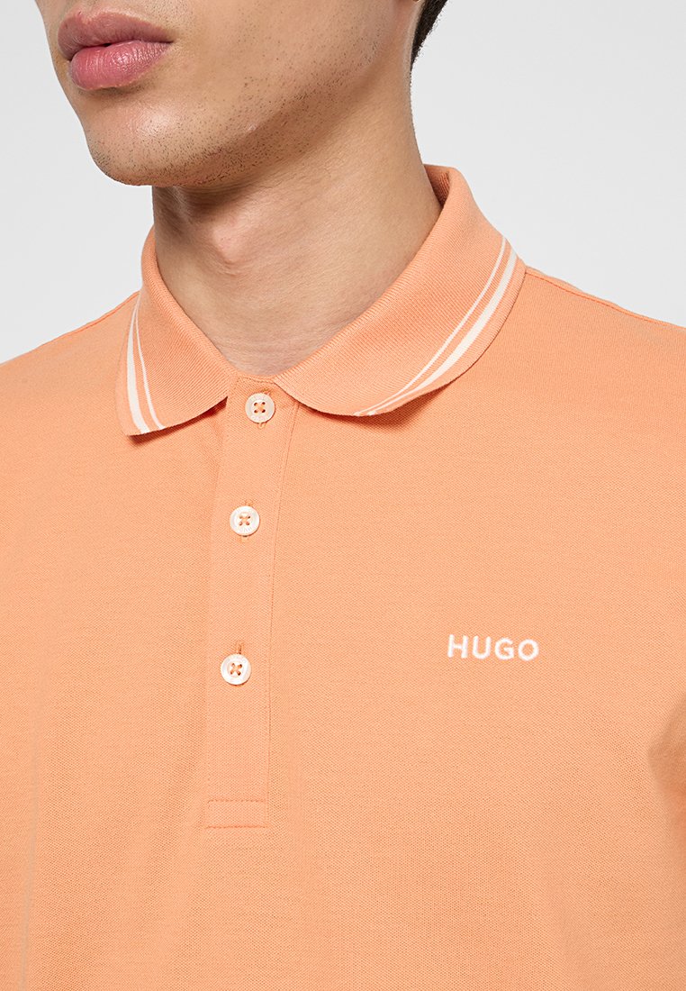 Jasnopomarańczowa koszulka polo wykonana z teksturowanej tkaniny, z klasycznym kołnierzykiem, zapięciem na trzy guziki i białym logo na piersi.