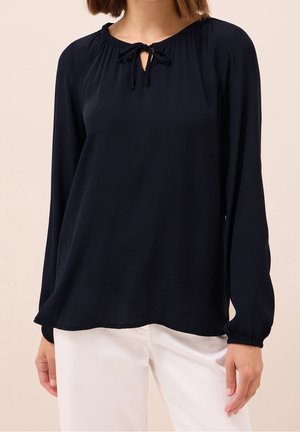 Femme portant un chemisier noir à manches longues avec un col noué, associé à un pantalon blanc sur un fond neutre.
