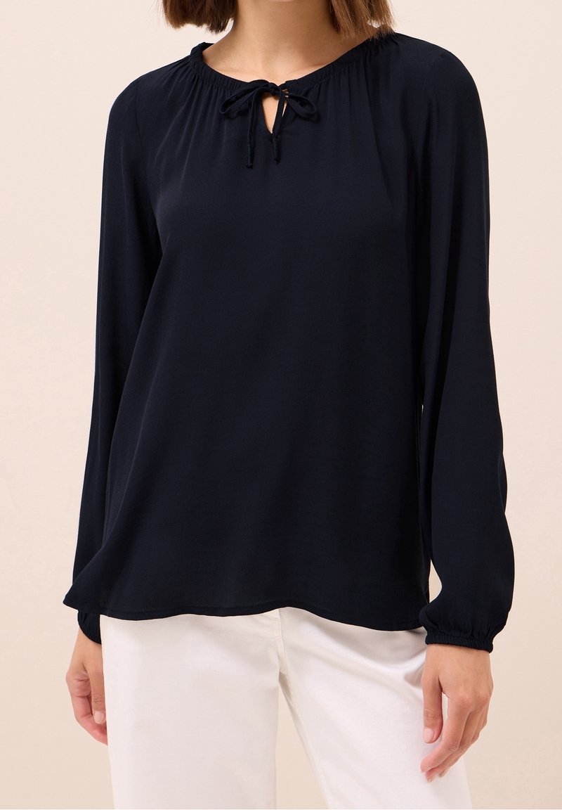 Femme portant un chemisier noir à manches longues avec un col noué, associé à un pantalon blanc sur un fond neutre.