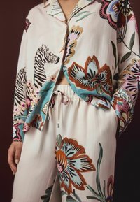 Woman wears cream-colored pajama set with large floral and zebra patterns, shirt partially open, hand resting in pants pocket.