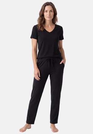 Femme debout pieds nus portant un haut noir à manches courtes en V et un pantalon noir à cordon sur un fond clair uni.
