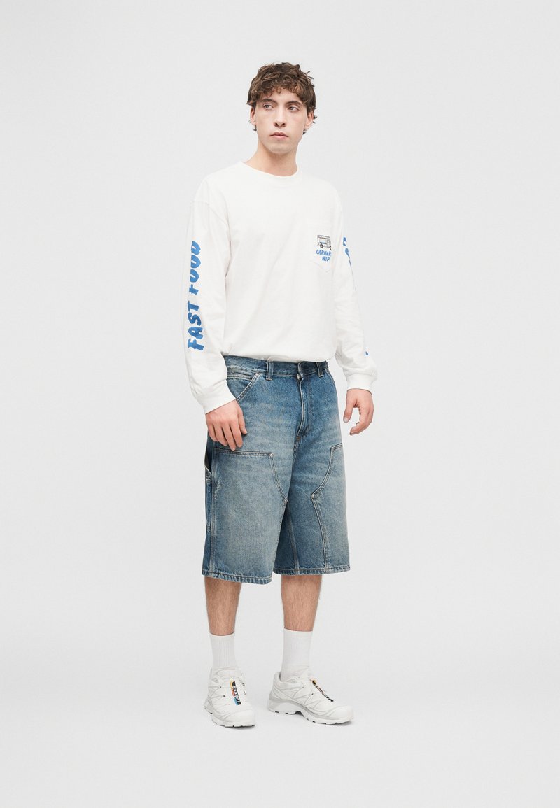 Hvid langærmet skjorte med blå tekst, kombineret med blå denimshorts. Fodtøj er hvide sneakers med mønstrede accenter.