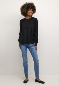 Schwarze Langarmbluse mit gerafften Details am Ausschnitt, kombiniert mit hellblauen Skinny-Jeans und schwarzen Flats, vor einem neutralen Hintergrund.