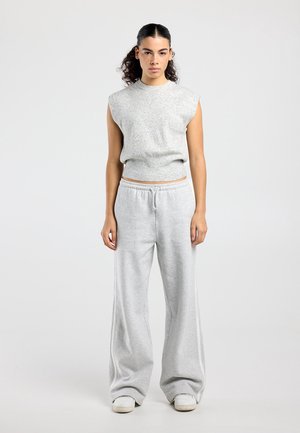 Vrouw met krullend haar in een hoge paardenstaart, gekleed in een mouwloos grijs shirt en losse grijze joggingbroek, staand tegen een effen lichte achtergrond.