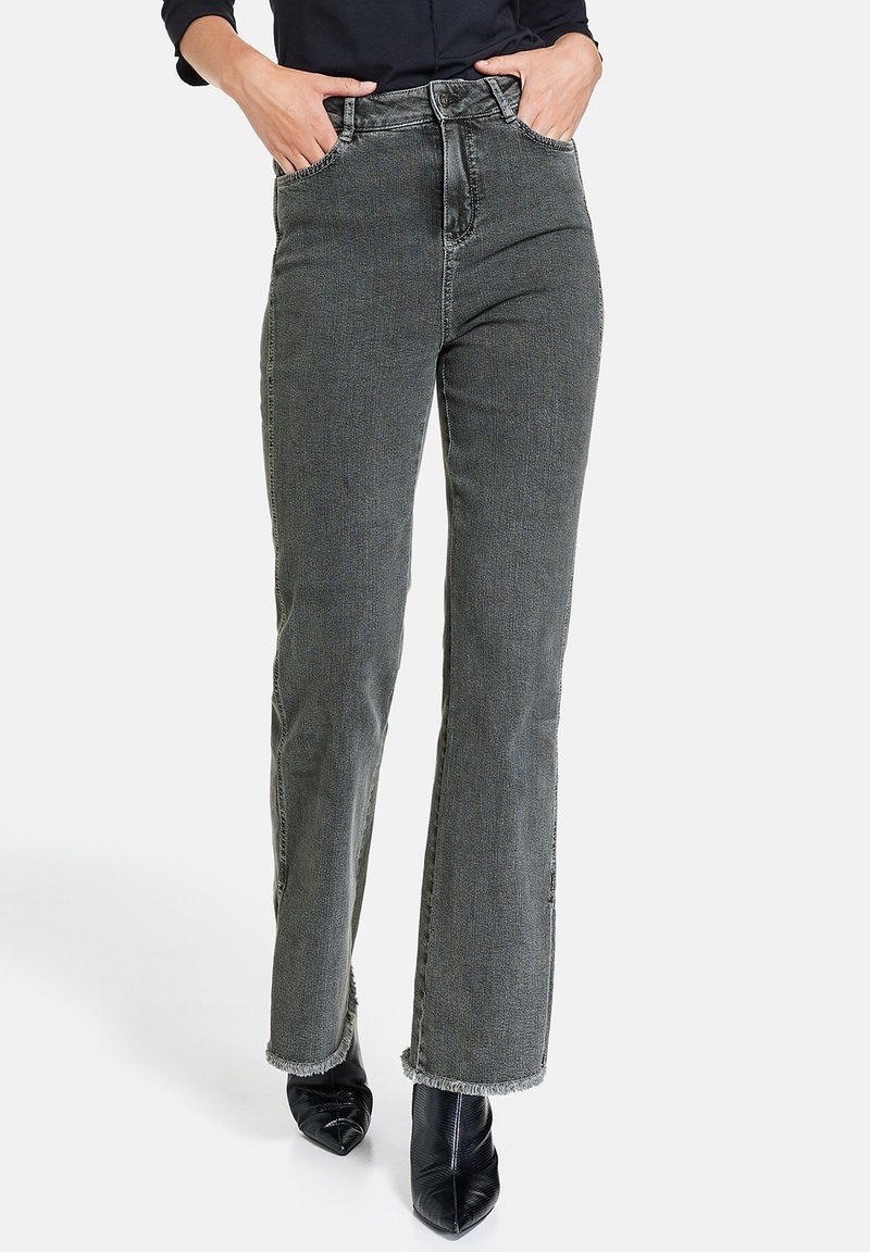 Femme portant un jean gris taille haute à jambes droites avec ourlets effilochés et bottines noires à bout pointu, mains dans les poches.