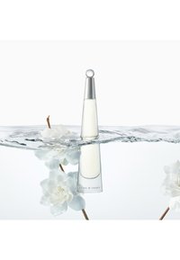 Una botella de perfume cónica y transparente con una tapa plateada, parcialmente sumergida en agua, rodeada de pétalos de flores blancas.