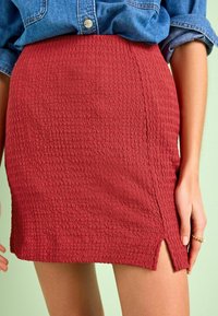 Gonna mini rossa, in tessuto strutturato con un motivo ondulato e una fessura laterale, realizzata in materiale elasticizzato. Abbinata a una camicia di denim annodata in vita.