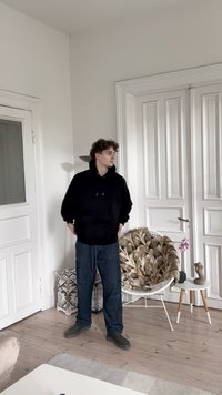 Uomo che indossa un hoodie nero con tasca a canguro e jeans blu navy, in piedi in una stanza con pareti bianche e pavimento in legno.