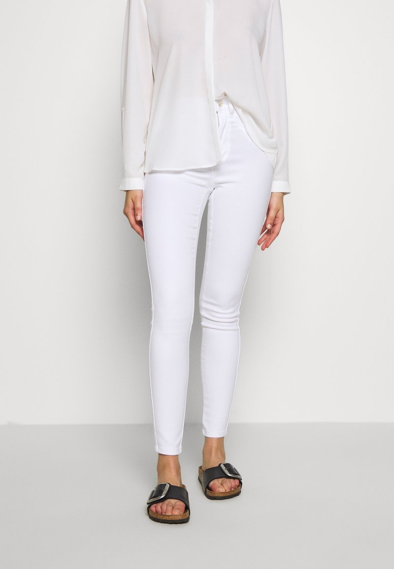 Jeans blancos ajustados con una textura suave, combinados con una camisa blanca clara y transparente de botones y sandalias planas negras con una hebilla grande.