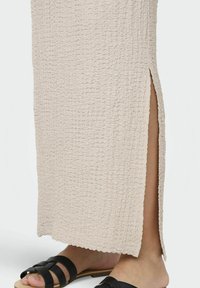Falda maxi texturizada beige con una abertura lateral, con un dobladillo en forma de ondas. Combinada con sandalias planas negras.