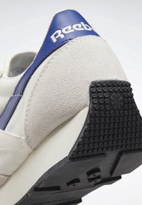 Vita och blå sportskor med ovandel i mocka, texturerad gummisula och prägling av logotyp på hälen. Solitt svart hälbas med grepp.