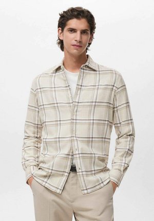 Camisa de cuadros en crema con líneas marrones, de mangas largas, corte holgado y con un bolsillo en el pecho. Llevada sobre una camiseta blanca.