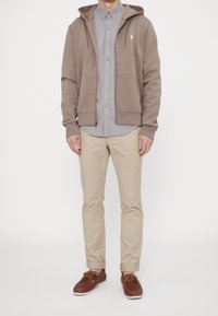 Felpa marrone con zip sopra una camicia abbottonata grigia chiaro, abbinata a pantaloni beige e scarpe marroni con lacci gialli. Design piatto e texture morbida.