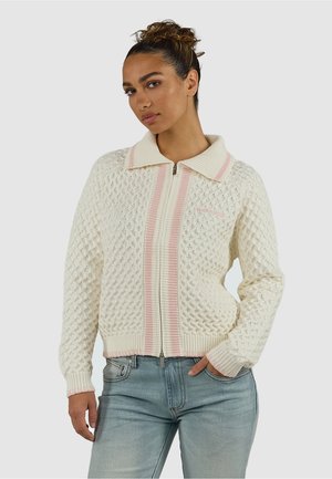 Vrouw die een crèmekleurige kabelgebreide sweater met rits en roze strepen draagt, staand met één hand in de lichtblauwe spijkerbroekzak tegen een effen achtergrond.