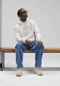 Camisola com capuz de cor creme com logótipo, jeans de ganga azul e sapatilhas claras. O modelo está sentado num banco de madeira contra um fundo neutro.