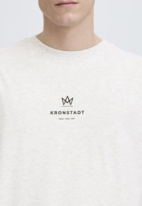 Camiseta de manga corta gris claro hecha de una tela suave, con un logo negro que presenta una corona y el texto "Kronstadt Casual Desde 1999" centrado debajo.