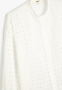 Hvid langærmet skjorte med knapper og rund krave samt firkantet udskåret mønster på stofsektioner, hængende mod en hvid baggrund.