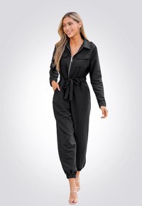 Combinaison noire avec un col, fermeture éclair à l'avant, deux poches sur la poitrine, poignets élastiques et détail de taille noué. Modèle portant des sandales à lanières.