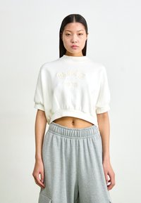 Sweatshirt corto branco com o logótipo felpudo "NIKE 1972", mangas curtas e um decote redondo, combinado com calças de moletom cinzentas com uma cintura elástica.