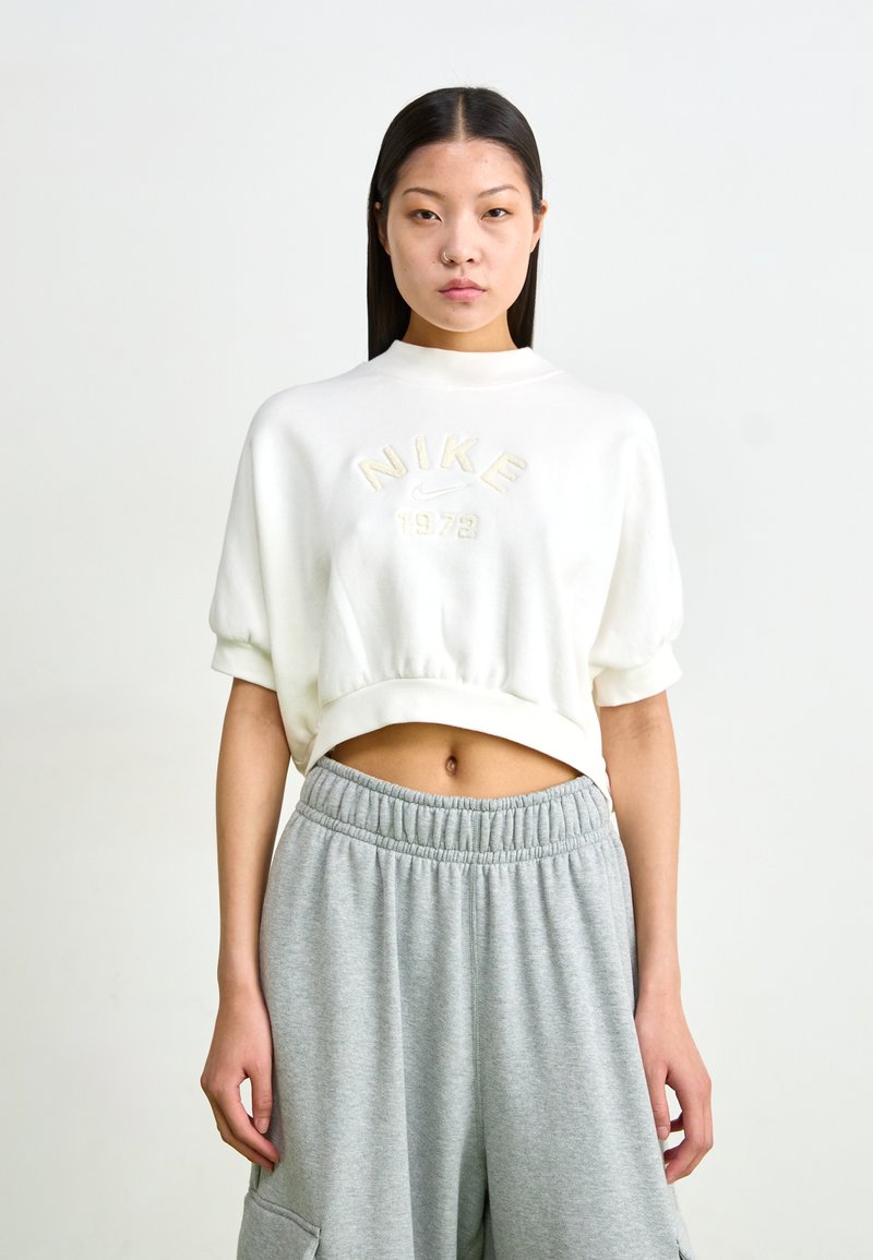 Sweatshirt corto branco com o logótipo felpudo "NIKE 1972", mangas curtas e um decote redondo, combinado com calças de moletom cinzentas com uma cintura elástica.