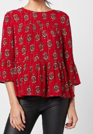 Woman wearing a red, floral-patterned, three-quarter sleeve blouse with ruffled hem paired with black leather pants.