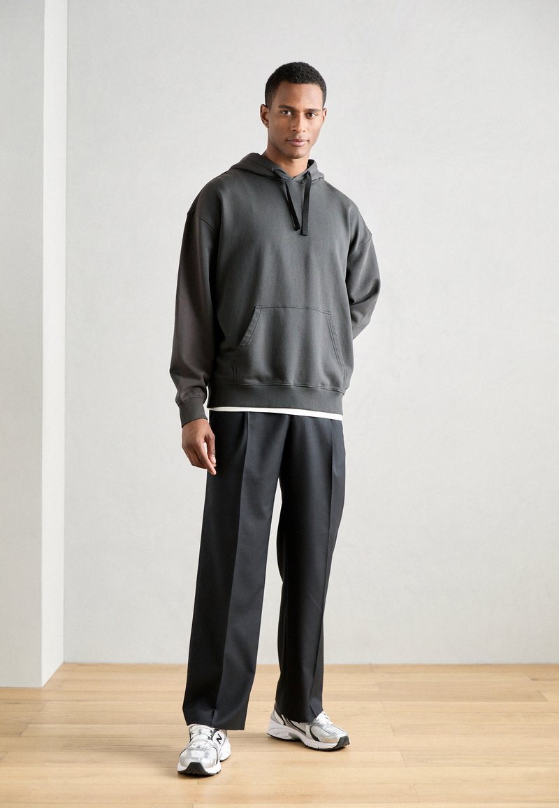 Donkergrijze oversized hoodie met een voorzak, gecombineerd met zwarte wijde broek en grijs-witte hardloopschoenen op een houten vloer.