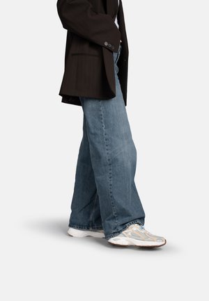 Persona che indossa jeans blu a gamba larga, sneakers bianche e beige e un blazer oversize scuro su uno sfondo semplice.