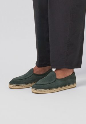 Persona que lleva alpargatas de ante verde oscuro con suela trenzada de yute y pantalones negros cortos contra un fondo claro.