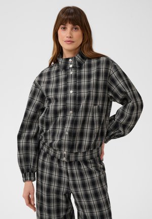 Femme portant une veste à carreaux noire et grise avec boutons-pression et un pantalon à carreaux assorti, debout avec une main sur la hanche sur fond blanc.