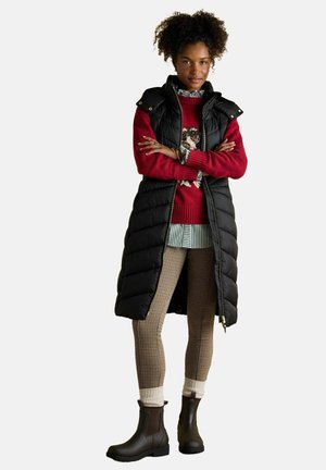 Schwarze lange Pufferweste über einem roten gemusterten Pullover, einem grün-weißen gestreiften Hemd und beigen karierten Leggings mit braunen Stiefeletten.