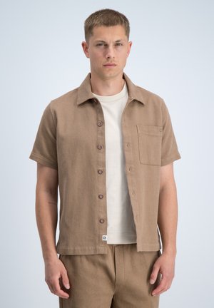 Man met een beige kortärmelig overhemd met knopen over een wit T-shirt, gecombineerd met bijpassende beige broek, staand tegen een effen achtergrond.