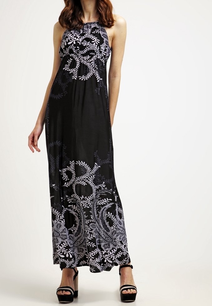 Femme portant une robe maxi noire sans manches avec des motifs floraux et cachemire blancs, associée à des sandales noires à talons hauts.