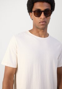 Tricou bej cu dungi, cu guler rotund și mâneci scurte. Model purtând ochelari de soare rotunzi închiși la culoare, evidențiind o textură netedă și moale.