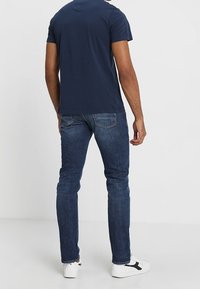 Camiseta de manga corta azul marino combinada con jeans oscuros de corte slim y zapatillas blancas con detalles en negro. Atuendo casual visto desde atrás.