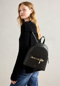 Sac à dos en cuir noir avec une forme arquée, doté d'une poche zippée à l'avant avec une doublure à imprimé léopard et des accents en métal doré.