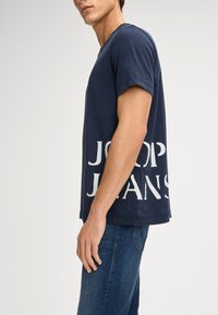 Navyblaue Baumwoll-T-Shirt mit kurzen Ärmeln und V-Ausschnitt, mit großem weißen "JOOP! JEANS"-Schriftzug an der Seite.