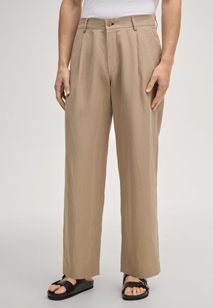 Uomo che indossa pantaloni beige a gamba larga con pieghe frontali, chiusura con bottoni, tasche laterali, camicia bianca infilata e sandali neri con fibbie.