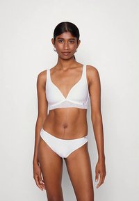 Γυναίκα με σκούρα μαλλιά φορώντας ένα λευκό, χωρίς ραφές σουτιέν και αντίστοιχα χαμηλής μέσης εσώρουχα, που στέκεται μπροστά σε μια απλή ανοιχτόχρωμη φόντο.