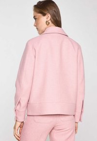 Veste en mélange de laine rose avec un col classique, épaules tombantes et fentes sur les manches. Texture lisse avec une coupe ajustée.
